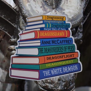 May include: A sticker featuring a stack of colorful books with titles like "Dragonsong," "Dragonflight," and "The White Dragon." The author's name, Anne McCaffrey, is also visible. The books have various colored spines and white pages.