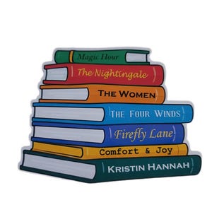 May include: A stack of seven books with colorful covers. Titles include "Magic Hour," "The Nightingale," "THE WOMEN," "THE FOUR WINDS," "Firefly Lane," "Comfort & Joy," and "KRISTIN HANNAH." The books are arranged in a neat pile.
