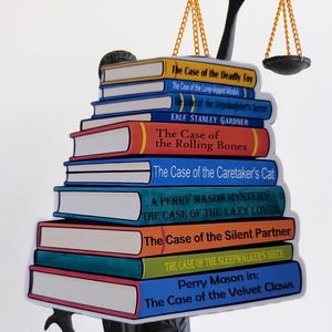 Op de afbeelding: Een stapel kleurrijke boeken met titels als "The Case of the Deadly Toy" en "The Case of the Rolling Bones", bekroond met een zwarte weegschaal. De boeken hebben witte pagina's en ruggen in verschillende kleuren, waaronder geel, blauw, rood en oranje.