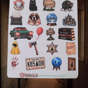 Puede incluir: Una hoja de pegatinas con varias ilustraciones de temática de terror que incluyen un payaso, un gato negro, una rata, una máquina de escribir, un perro, una bola de nieve, una casa, un coche verde, un globo rojo, un cartel y varios libros.