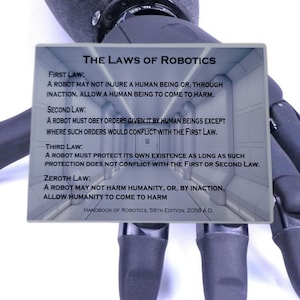 Op de afbeelding: Een rechthoekig metalen bord met de tekst The Laws of Robotics, met de eerste, tweede, derde en nulde wet, en de vermelding Handbook of Robotics, 56th Edition, 2058 A.D. Het bord is geplaatst voor een zwarte robotische hand.