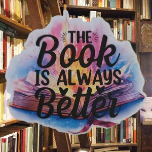 Può includere: Un'insegna colorata con la scritta "The Book Is Always Better" in corsivo nero, ambientata su un'illustrazione di un libro aperto con effetto acquerello. Lo sfondo mostra una libreria in legno piena di libri.