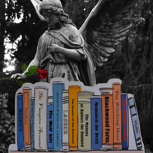 Puede incluir: Una estantería de libros de Anne Rice, con títulos como "The Vampire Armand", "The Wolf Gift", "Interview with the Vampire", "Lasher", "The Queen of the Damned", "The Mummy or Ramses the Damned", "Memnoch the Devil", "Blackwood Farm", "The Witching Hour", "The Vampire Lestat" y "Violin".