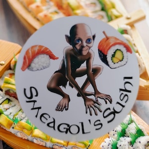 May include: A wooden boat filled with various sushi rolls. A white circle sticker with a cartoon character and the text "Sheagol's Sushi" is placed on top of the boat.