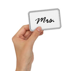 May include: A white rectangular patch with the word "Mrs." in black script. The patch has a grey stitched border and is held by a hand against a white background.
