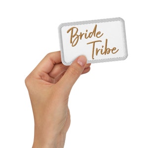 May include: White fabric patch with a gold embroidered design that says "Bride Tribe".