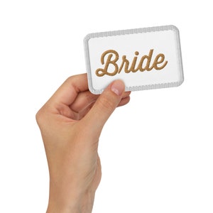 May include: A white rectangular patch with the word "Bride" embroidered in a gold-colored script. The patch has a gray stitched border and is held in a hand against a white background. The patch is a wedding accessory.