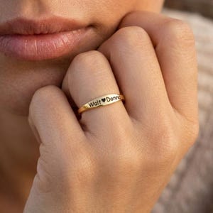 May include: A gold-toned band ring featuring a flat, polished surface engraved with the text "Walt" followed by a small heart symbol and the name "Donna" in a clear, dark, sans-serif font, worn on a finger.