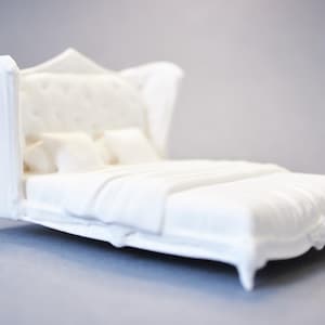 May include: A miniature white bed with a tufted headboard and wingback sides. The bed is covered with a white blanket and pillows. The bed frame has decorative detailing and sits on four small legs. The overall theme is bedroom decor.