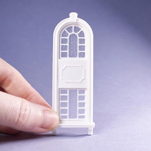May include: A white miniature architectural window with an arched top and rectangular panes. The window has a decorative panel in the center and is held by a hand against a purple background. The window is likely for a dollhouse or model.