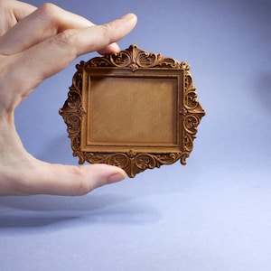 May include: A small, ornate, rectangular picture frame held by a hand. The frame is a warm brown colour with intricate scrollwork and decorative detailing. The background is a soft, muted blue.