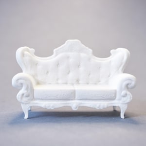 Op de afbeelding: Een miniatuur witte bank met een klassiek, sierlijk ontwerp. De bank heeft een gecapitonneerde rugleuning en zitting, gekrulde armleuningen en decoratieve poten. De algehele stijl doet denken aan antieke meubels.