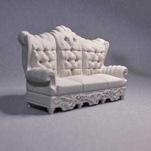 May include: A white miniature sofa with a tufted back and seat. The sofa has ornate carvings along the base and arms. The sofa is a small scale model, likely for a dollhouse or decorative display.
