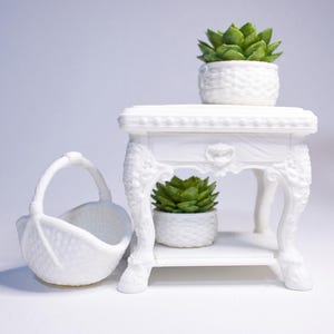May include: A white, ornate side table with a small succulent plant in a white pot on top and another on the lower shelf. A white basket is on the left. The table and basket have a textured surface.