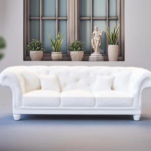 Puede incluir: Un sofá blanco de tres plazas con respaldo capitoné y brazos enrollados. El sofá tiene tres cojines blancos y está frente a una ventana con plantas en macetas y una estatua. La habitación está decorada con plantas de interior.