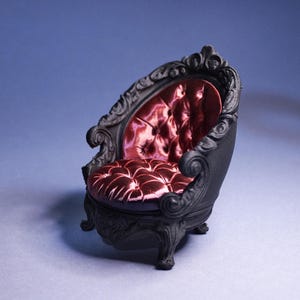 Può includere: Una poltrona nera in miniatura con dettagli ornati e una seduta in velluto rosa-rossastro trapuntato. La sedia ha braccioli e gambe curve e uno schienale decorativo. Il design generale ricorda i mobili antichi.