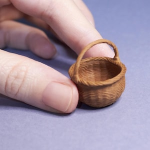 Könnte beinhalten: Ein winziger, brauner Korb mit Henkel. Der Korb wird zwischen Fingern vor einem hellvioletten Hintergrund gehalten. Die Textur und Form des Korbes sind deutlich erkennbar.