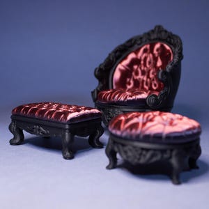 Puede incluir: Juego de muebles en miniatura negros con asiento de terciopelo burdeos acolchado. El conjunto incluye una silla y dos otomanas, todas con detalles ornamentados y enrollados. El fondo es azul liso.