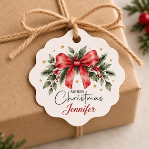 May include: A white scalloped gift tag featuring a watercolor illustration of a red bow, green holly leaves, and red berries. The tag includes gold stars and the black text Merry Christmas with the red script name Jennifer printed below it.