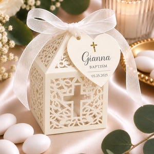 May include: A cream-colored laser-cut favor box with a cross cutout on each side, tied with a white sheer ribbon. A heart-shaped tag attached to the ribbon features a gold cross, the name Gianna, the word BAPTISM, and the date 05.25.2025.