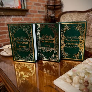 May include: Three hardcover books with gold lettering on a green background. The titles are "The Cruel Prince", "The Wicked King", and "The Queen of Nothing".