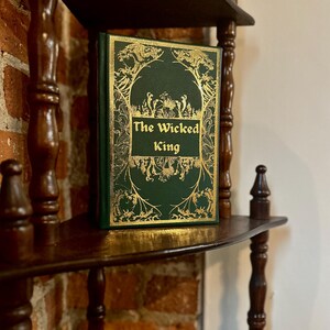 May include: A hardcover book titled "The Wicked King" with a green cover and gold decorative accents. The book is displayed on a dark wooden shelf, showcasing its ornate design and title.