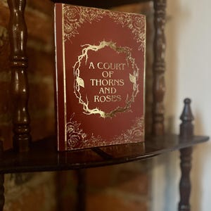 May include: A red book with gold lettering on the cover. The title is "A Court of Thorns and Roses".