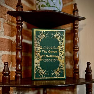 May include: A green book with gold leaf detailing on a wooden shelf. The title of the book is "The Queen of Nothing".