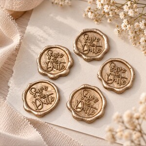 May include: Five circular gold wax seals with irregular, scalloped edges. Each seal features the embossed text "Save the Date" in a cursive script. The seals have a metallic finish and are arranged on a light-colored, textured paper surface.