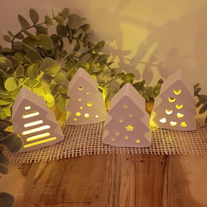 Könnte beinhalten: Vier weiße Keramik-Weihnachtsbaum-Kerzenhalter mit ausgeschnittenen Designs. Jeder Baum hat ein anderes Muster, darunter Linien, Kreise und Sterne. Die Kerzenhalter stehen auf einer Holzoberfläche mit einer gewebten Matte und Grünzeug im Hintergrund.