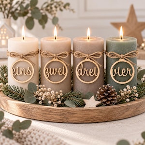 May include: Four cylindrical pillar candles in shades of cream, light brown, taupe, and sage green. Each candle features a lit wick and a circular wooden tag tied with twine, displaying the German words eins, zwei, drei, and vier respectively.