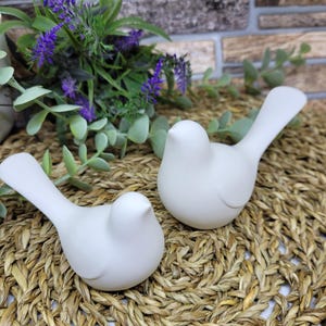 May include: Two white ceramic birds with long tails, sitting on a woven straw mat. The birds are facing different directions.