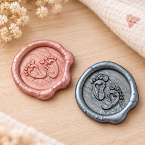 May include: Two circular wax seals, one in a metallic rose gold finish and the other in a metallic slate gray finish. Each seal features an embossed design of two small baby footprints with heart-shaped soles centered within the circular border.