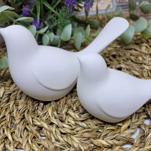 May include: Two white ceramic bird figurines, one with its head turned to the side, sitting on a woven straw mat.