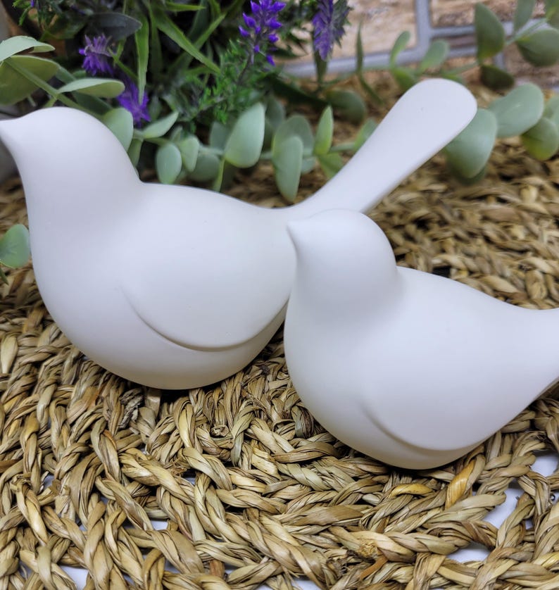 May include: Two white ceramic bird figurines, one with its head turned to the side, sitting on a woven straw mat.