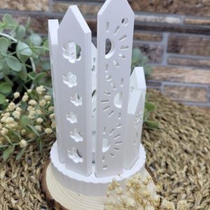 May include: White wooden candle holder with a geometric design featuring cutouts of suns, moons, and clouds. The holder is on a wooden base.