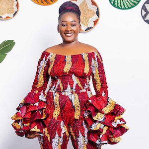 May include: A vibrant, off-the-shoulder jumpsuit in red, yellow, and white patterns. The jumpsuit features long sleeves with ruffled cuffs and flared legs. Decorative woven wall hangings in various colors and patterns are in the background.