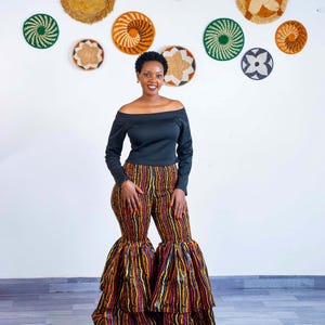 May include: A woman wearing a black off-the-shoulder top and flared pants with vertical stripes in shades of orange, yellow, and purple. Woven wall decor in various sizes and patterns hangs on the white wall behind her.