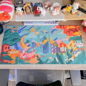 May include: A colorful desk mat featuring a whimsical illustration of a fantasy landscape with cartoonish characters, trees, and buildings. The mat has a blue background with orange, green, and yellow accents. Text on the mat reads "POYO islands" and "WA-WA!"