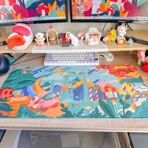 May include: A colorful desk setup featuring a large, rectangular mousepad with a whimsical illustration. The mousepad has a vibrant design with cartoonish elements, including a pig wearing goggles and the word "POYO". Various figurines and a keyboard are also visible.