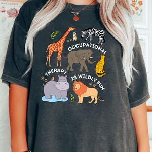 May include: Dark grey t-shirt with a colorful graphic featuring cartoon animals including a giraffe, zebra, elephant, cheetah, hippo, and lion. The text "OCCUPATIONAL THERAPY IS WILDLY FUN" is printed on the shirt.