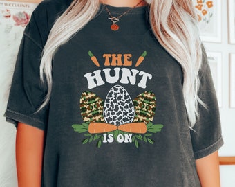 May include: A dark gray t-shirt with the text "The Hunt Is On" in white. The text is surrounded by a graphic of three Easter eggs, one with a camouflage pattern, one with a leopard print, and one with a white and black speckled pattern. The eggs are surrounded by green leaves and orange carrots.