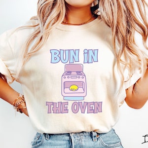 May include: A white t-shirt with the text "Bun in the Oven" in pink and blue lettering. A cartoon pink oven with a yellow bun inside is pictured below the text.