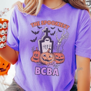 May include: Purple t-shirt with a spooky Halloween design. The design features a tombstone with the words "RIP" on it, a skeleton hand, and three pumpkins. The text "The Spookiest BCBA" is printed on the shirt.