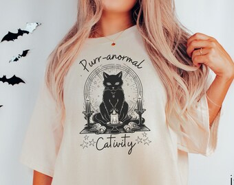 May include: A white t-shirt with a black cat illustration and the text "Purr-anormal Cativity" printed on the front.
