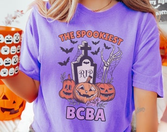 May include: Purple t-shirt with a spooky Halloween design. The design features a tombstone with the words "RIP" on it, a skeleton hand, and three pumpkins. The text "The Spookiest BCBA" is printed on the shirt.