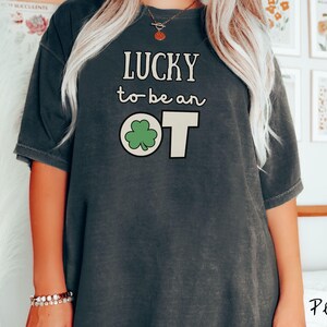 May include: Dark gray t-shirt with the text "LUCKY to be an OT" in white. The "O" in "OT" is replaced with a green shamrock. The shirt is a casual style.