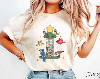 May include: Ivory-colored t-shirt featuring a whimsical bird feeder design. The graphic includes a star, colorful birds, and a bird feeder filled with seeds. The design is surrounded by small stars, creating a cheerful and nature-inspired aesthetic.