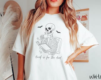 May include: White t-shirt with a black skeleton graphic reading a book and drinking coffee, with two bats above. The text "decaf is for the dead" is printed below the skeleton. The shirt is a casual, oversized fit.