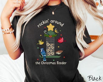 May include: Black t-shirt with a colorful graphic of a Christmas tree-shaped bird feeder. The design includes various birds, a star, and the text "rockin' around the Christmas Feeder."